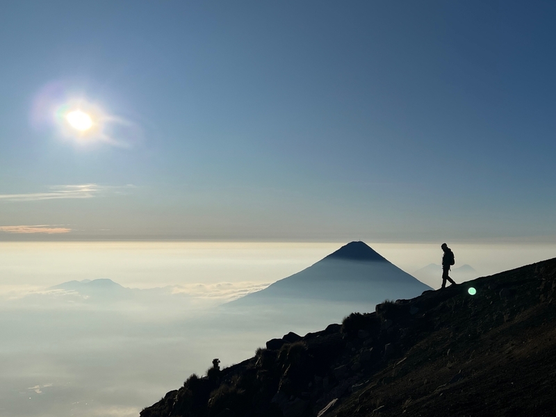 Hiking-with-a-silhouette-of-a-hiker-against-the-backdrop-of-a-volcano-towering-over-the-clouds-in-the-background.-Voldaco-de-Fuego-Acatenango-antigua-Guatemala.