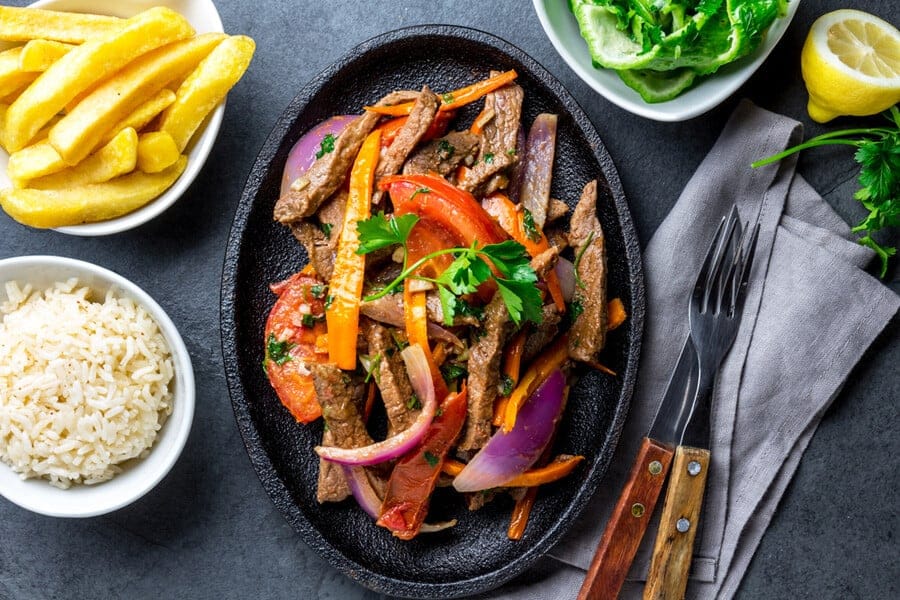 Peruvian-dish-Lomo-saltado-beef-tenderloin-with-purple-onion-yellow-chili-tomatoes-served-on-black-plate-with-french-fries-and-rice.-Top